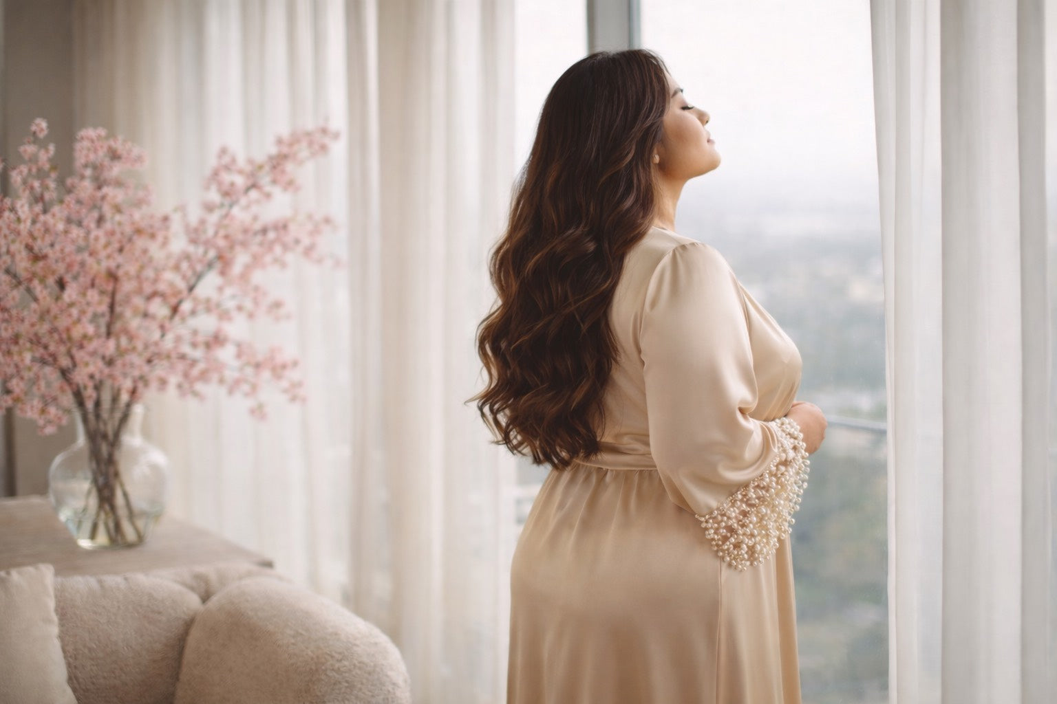 Femme élégante de profil, debout près d’une grande fenêtre, respirant calmement, vêtue d’un ensemble satin beige doré, dans un intérieur lumineux et apaisant aux tons Hazellia, symbole de bien-être et de reconnexion au corps féminin.