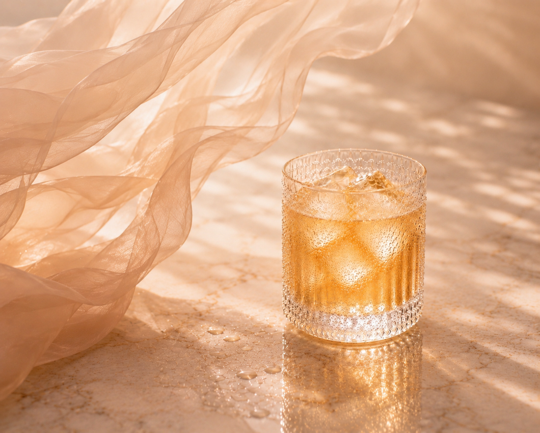GlowTox Hazellia – infusion glacée dorée dans un verre cristal premium avec glaçons translucides, condensation naturelle, zeste de citron vert et voile rose poudré couture sur marbre luxueux.


