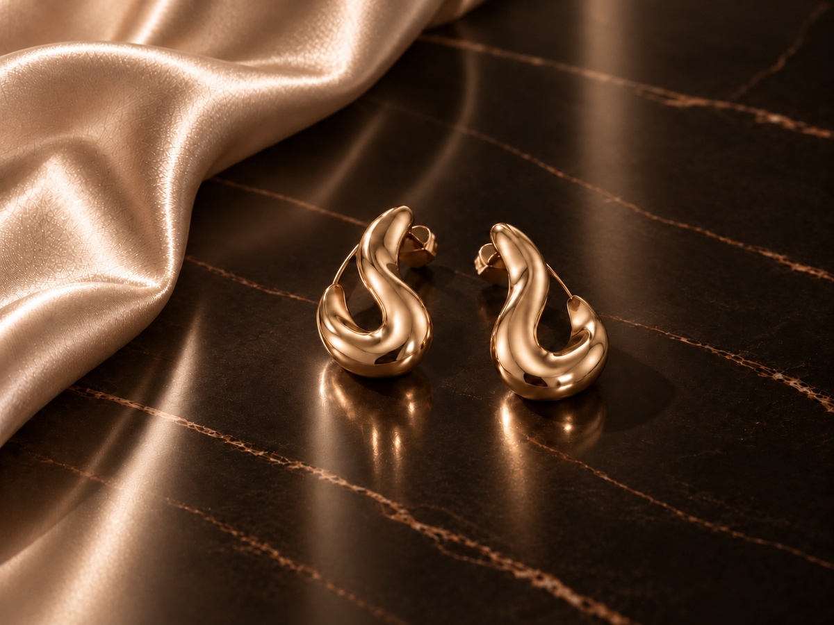 Deux boucles d’oreilles dorées sculpturales sur marbre espresso brillant et satin champagne, univers élégant de la rubrique Saison Dorée du Magazine Hazellia.
